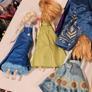 May include: A collection of dolls with long hair and vibrant dresses. One doll is in a blue dress with a cape, another in a green dress, and a third doll is wearing a blue skirt with a sunflower design. The dolls are inspired by a well-known animated film.