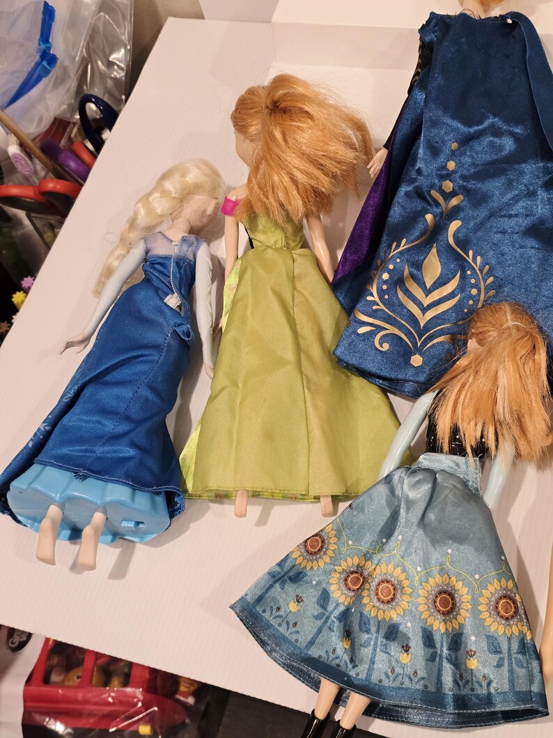 May include: A collection of dolls with long hair and vibrant dresses. One doll is in a blue dress with a cape, another in a green dress, and a third doll is wearing a blue skirt with a sunflower design. The dolls are inspired by a well-known animated film.