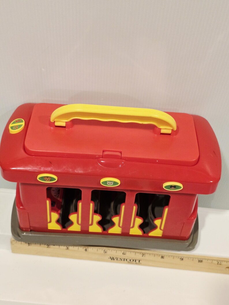 K&ouml;nnte beinhalten: Roter und gelber Spielzeugbus mit gelbem Griff oben. Das Spielzeug hat drei &Ouml;ffnungen vorne und drei runde Kn&ouml;pfe mit gr&uuml;n-gelben Designs. Ein Lineal ist vor dem Spielzeug platziert.