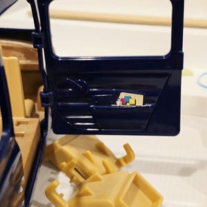 May include: A navy blue toy car door is open, revealing a tan interior. Two tan plastic pieces are below the door. A small colorful graphic is visible inside the door.