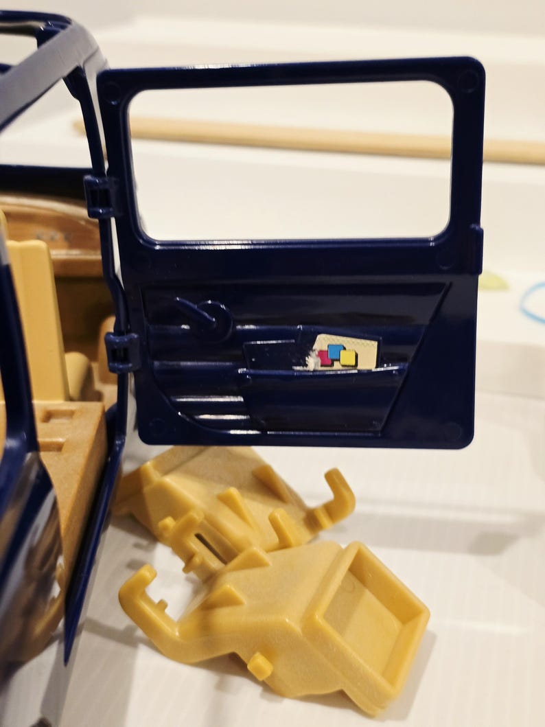 May include: A navy blue toy car door is open, revealing a tan interior. Two tan plastic pieces are below the door. A small colorful graphic is visible inside the door.