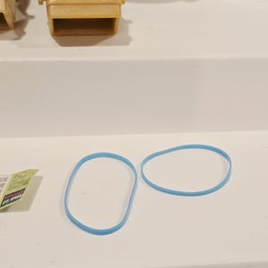 May include: Two light blue rubber bands are displayed on a white surface. The bands are oval-shaped and appear to be made of a flexible material. A small, tan plastic chair is also visible in the background.