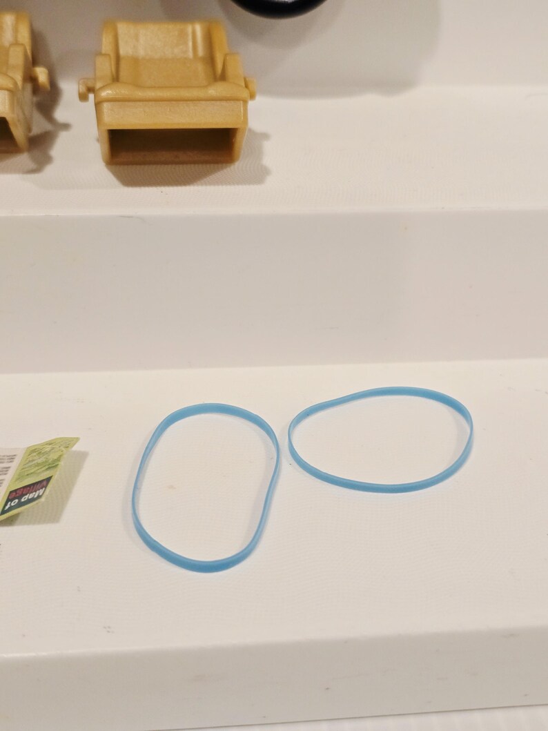 May include: Two light blue rubber bands are displayed on a white surface. The bands are oval-shaped and appear to be made of a flexible material. A small, tan plastic chair is also visible in the background.