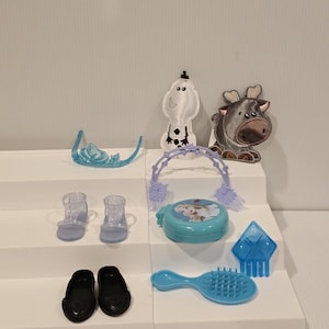 K&ouml;nnte beinhalten: Eine Sammlung von Kinderspielzeug und Zubeh&ouml;r, darunter ein blauer Diadem, transparente Abs&auml;tze, schwarze Ballerinas, ein blauer Kompaktpuder mit Charakterbild, eine blaue Haarb&uuml;rste und Figuren eines Schneemanns und eines Rentiers.