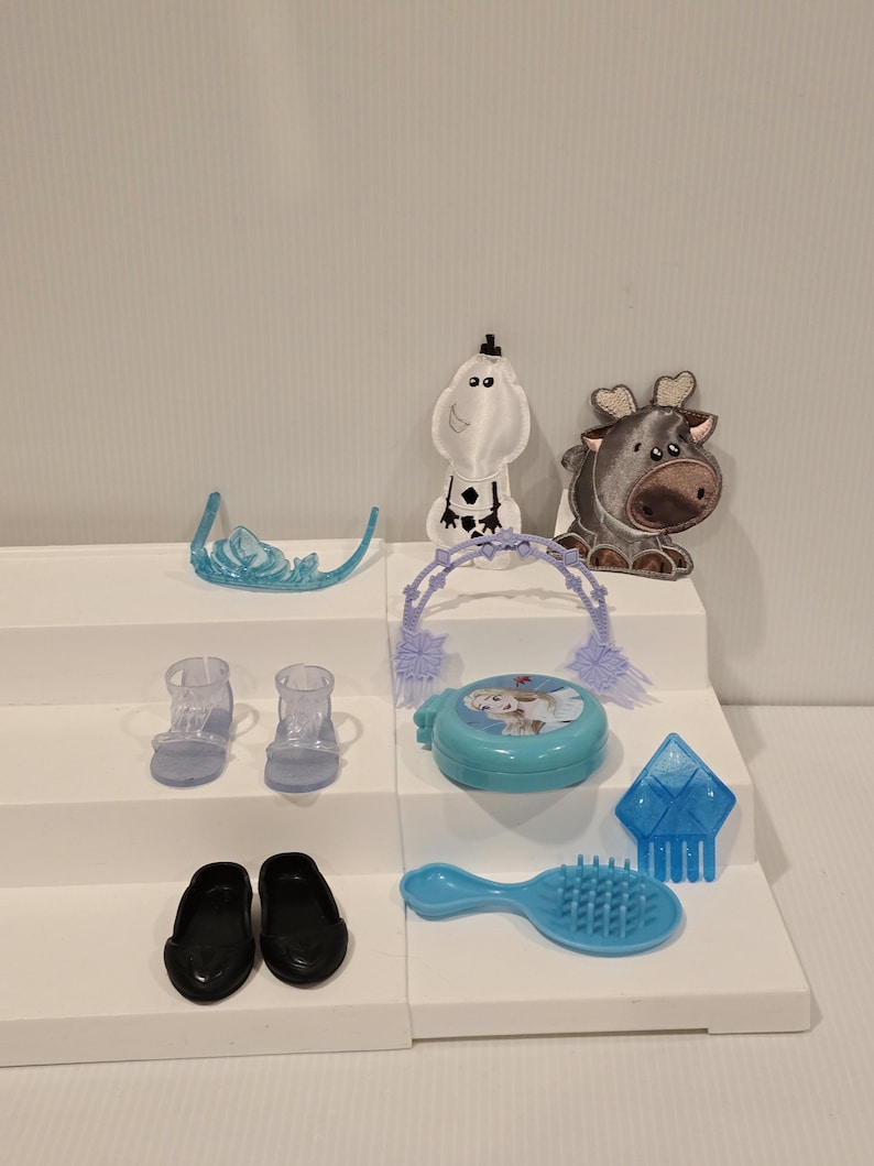 K&ouml;nnte beinhalten: Eine Sammlung von Kinderspielzeug und Zubeh&ouml;r, darunter ein blauer Diadem, transparente Abs&auml;tze, schwarze Ballerinas, ein blauer Kompaktpuder mit Charakterbild, eine blaue Haarb&uuml;rste und Figuren eines Schneemanns und eines Rentiers.