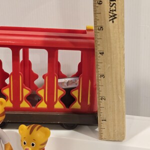 May include: A red and yellow toy train car with a brown base. The train car has a cage-like design with yellow starburst accents. A wooden ruler is next to the train car. A Daniel Tiger figurine is in the foreground.