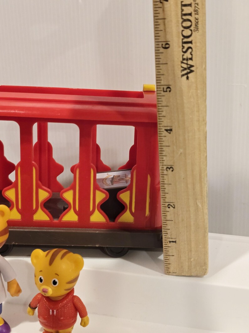 May include: A red and yellow toy train car with a brown base. The train car has a cage-like design with yellow starburst accents. A wooden ruler is next to the train car. A Daniel Tiger figurine is in the foreground.