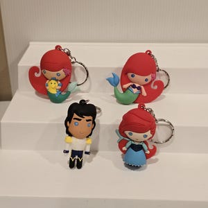 May include: Four character keychains featuring characters from a popular animated film. The keychains include two mermaids with red hair and green tails, a prince in a white and gold uniform, and a mermaid in a blue dress.