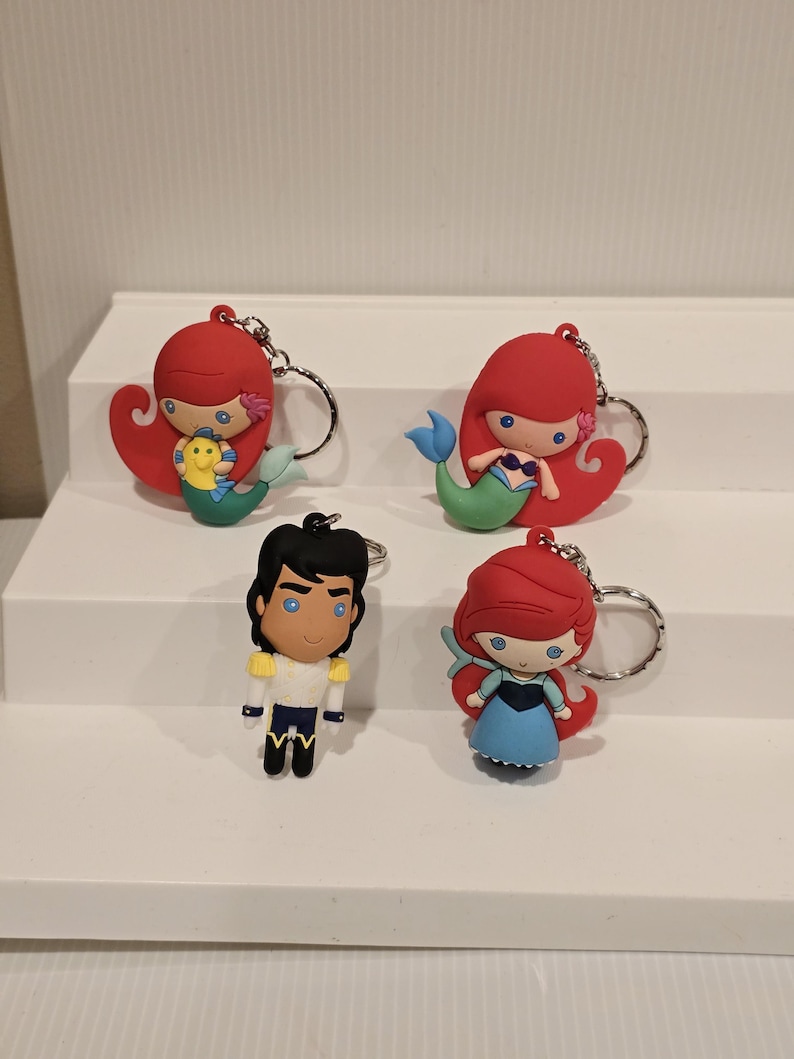 May include: Four character keychains featuring characters from a popular animated film. The keychains include two mermaids with red hair and green tails, a prince in a white and gold uniform, and a mermaid in a blue dress.