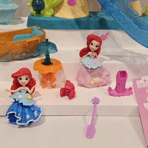 K&ouml;nnte beinhalten: Ariel-Spielset mit Miniaturfiguren von Ariel, einer Krabbe und Zubeh&ouml;r. Das Set enth&auml;lt ein Boot, einen Tisch mit einer Lampe und eine Rutsche. Die Figuren sind in verschiedenen Posen und tragen farbenfrohe Outfits. Der Hintergrund umfasst ein Schloss.