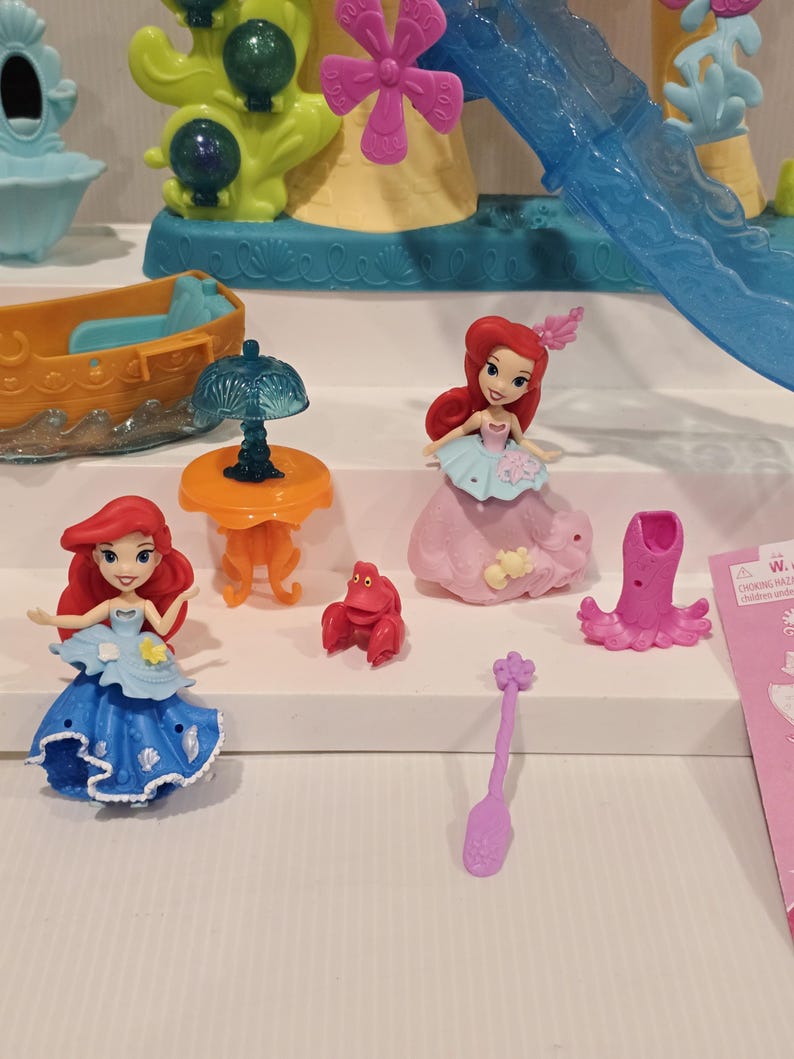 K&ouml;nnte beinhalten: Ariel-Spielset mit Miniaturfiguren von Ariel, einer Krabbe und Zubeh&ouml;r. Das Set enth&auml;lt ein Boot, einen Tisch mit einer Lampe und eine Rutsche. Die Figuren sind in verschiedenen Posen und tragen farbenfrohe Outfits. Der Hintergrund umfasst ein Schloss.