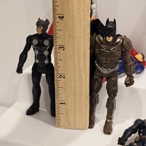 May include: A wooden ruler stands next to two action figures: Thor and Batman. The Batman figure is approximately 3 inches tall. Thor is black with silver accents. Batman is brown with a black cape. Other figures are partially visible.