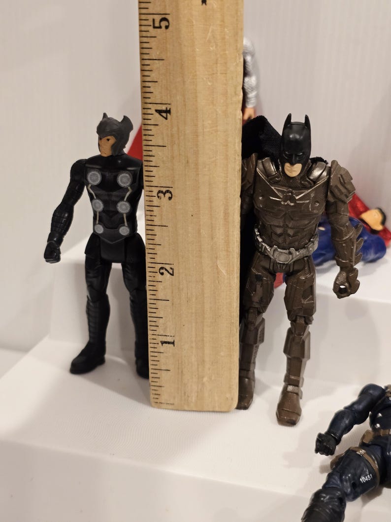 May include: A wooden ruler stands next to two action figures: Thor and Batman. The Batman figure is approximately 3 inches tall. Thor is black with silver accents. Batman is brown with a black cape. Other figures are partially visible.