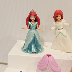 K&ouml;nnte beinhalten: Mehrere Ariel-Figuren werden auf einer wei&szlig;en Oberfl&auml;che pr&auml;sentiert. Eine Figur tr&auml;gt ein t&uuml;rkisfarbenes Kleid und eine silberne Krone. Eine andere tr&auml;gt ein wei&szlig;es Kleid. Eine gelbe Fischfigur ist ebenfalls zu sehen, zusammen mit einem rosa Kleid.