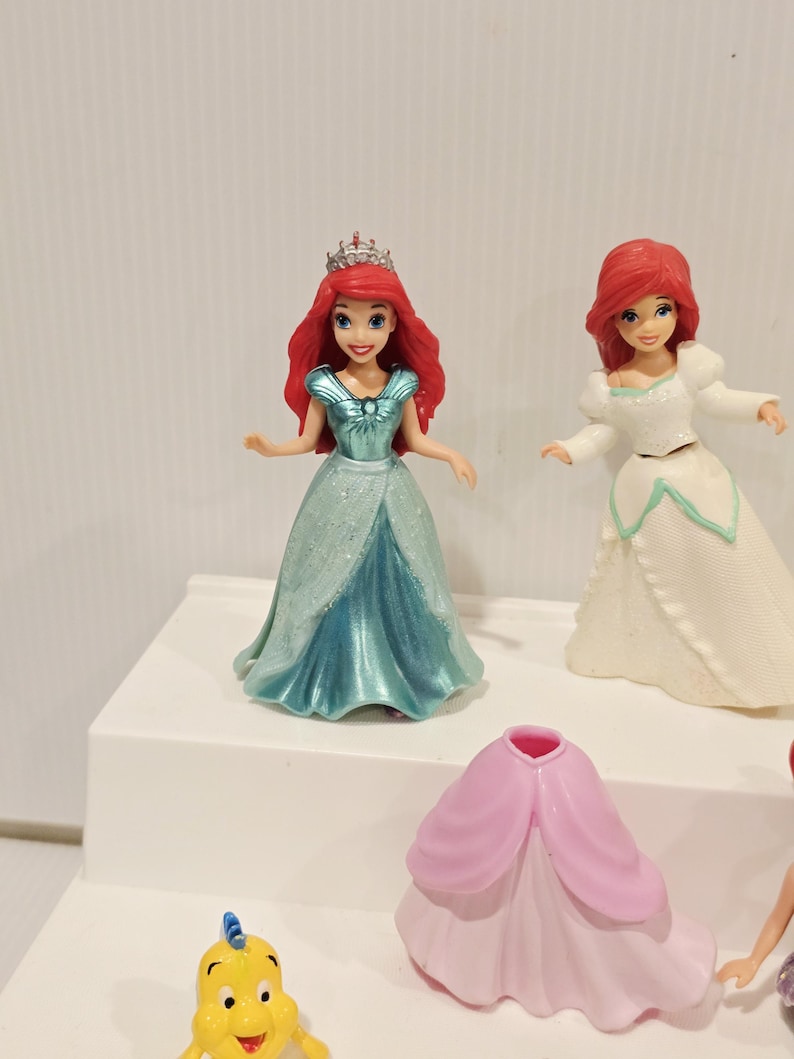 K&ouml;nnte beinhalten: Mehrere Ariel-Figuren werden auf einer wei&szlig;en Oberfl&auml;che pr&auml;sentiert. Eine Figur tr&auml;gt ein t&uuml;rkisfarbenes Kleid und eine silberne Krone. Eine andere tr&auml;gt ein wei&szlig;es Kleid. Eine gelbe Fischfigur ist ebenfalls zu sehen, zusammen mit einem rosa Kleid.
