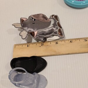 K&ouml;nnte beinhalten: Ein grauer und brauner Haarklammer in Form eines Nilpferds, ein schwarzer Schuh und eine transparente Sandale werden gezeigt. Ein Holzlineal befindet sich neben den Artikeln. Eine blaue Haarb&uuml;rste mit lila Borsten ist im Hintergrund.