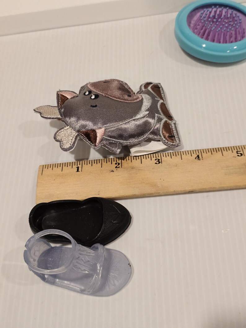 K&ouml;nnte beinhalten: Ein grauer und brauner Haarklammer in Form eines Nilpferds, ein schwarzer Schuh und eine transparente Sandale werden gezeigt. Ein Holzlineal befindet sich neben den Artikeln. Eine blaue Haarb&uuml;rste mit lila Borsten ist im Hintergrund.