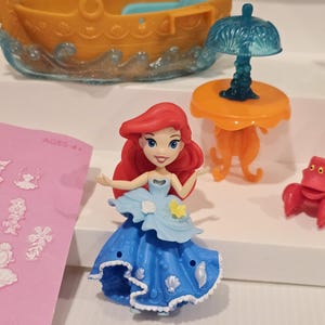 K&ouml;nnte beinhalten: Ariel-Figur aus dem Disney-Film Arielle, die Meerjungfrau. Die Puppe hat rotes Haar und ein blaues Kleid. Weitere Spielzeuge sind ein Boot, ein Tisch mit einer Lampe und eine rote Krabbe.