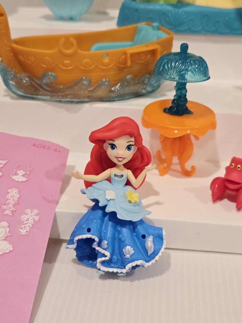 K&ouml;nnte beinhalten: Ariel-Figur aus dem Disney-Film Arielle, die Meerjungfrau. Die Puppe hat rotes Haar und ein blaues Kleid. Weitere Spielzeuge sind ein Boot, ein Tisch mit einer Lampe und eine rote Krabbe.