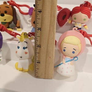 May include: A selection of character keyrings, featuring a dog, a unicorn, and a doll. The keyrings are attached to red cords. A wooden ruler is placed next to the keyrings, measuring approximately 12.7 cm.