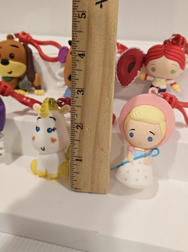 May include: A selection of character keyrings, featuring a dog, a unicorn, and a doll. The keyrings are attached to red cords. A wooden ruler is placed next to the keyrings, measuring approximately 12.7 cm.