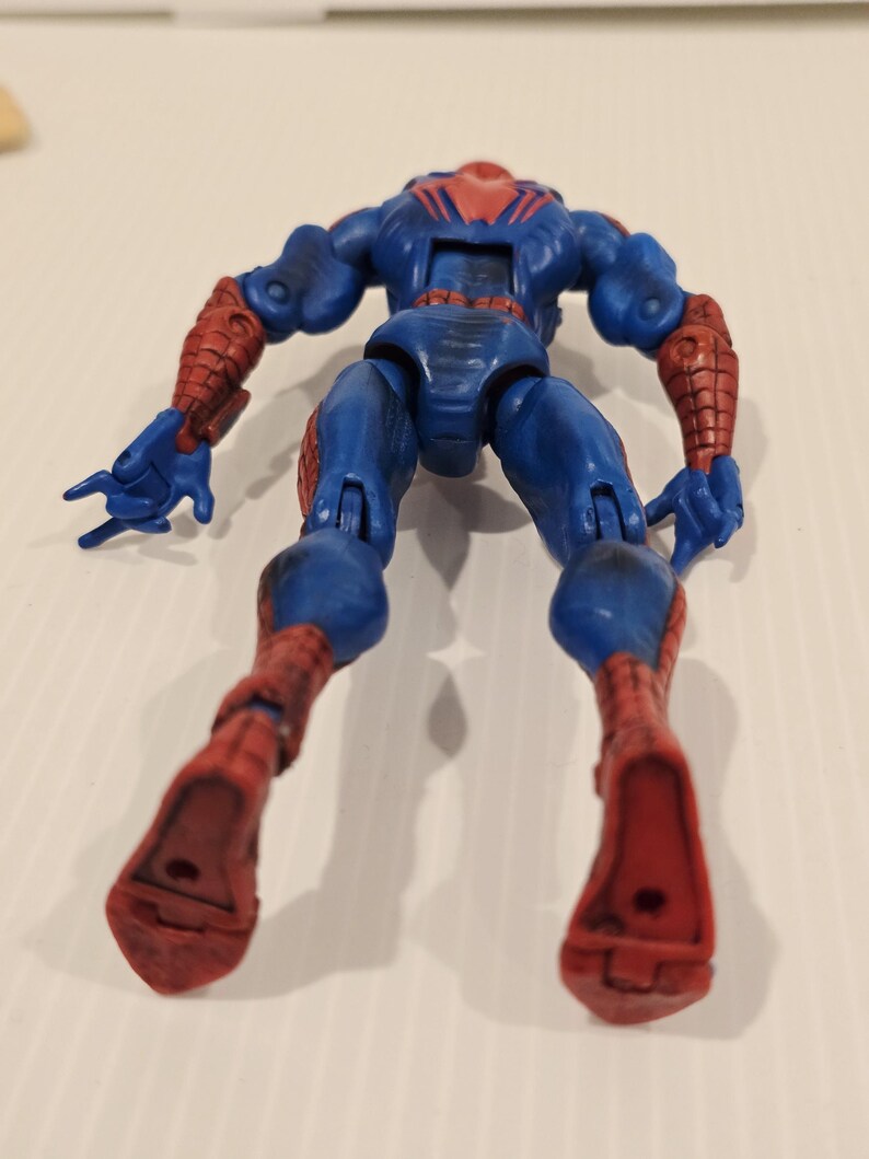 Pode incluir: Figura de a&ccedil;&atilde;o azul e vermelha de um super-her&oacute;i, vista de tr&aacute;s. A figura tem um corpo azul com detalhes vermelhos nos bra&ccedil;os e pernas. A cabe&ccedil;a &eacute; vermelha com um desenho de teia de aranha preta. As m&atilde;os s&atilde;o azuis.