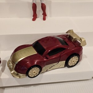 May include: A red and gold toy car with a spoiler. The car has gold accents on the hood, sides, and spoiler. The wheels are beige. Other action figures are visible in the background.
