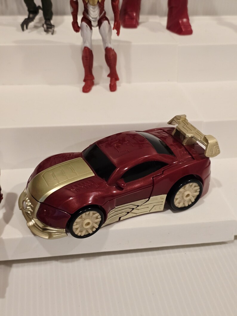May include: A red and gold toy car with a spoiler. The car has gold accents on the hood, sides, and spoiler. The wheels are beige. Other action figures are visible in the background.