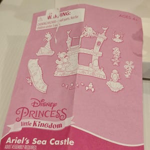 K&ouml;nnte beinhalten: Rosa Anleitung f&uuml;r das Disney Princess Little Kingdom Ariel's Sea Castle Spielset. Die Anleitung zeigt ein Schlossdesign und verschiedene Charakterillustrationen. Der Text enth&auml;lt eine Warnung vor Erstickungsgefahr und das Hasbro-Logo.