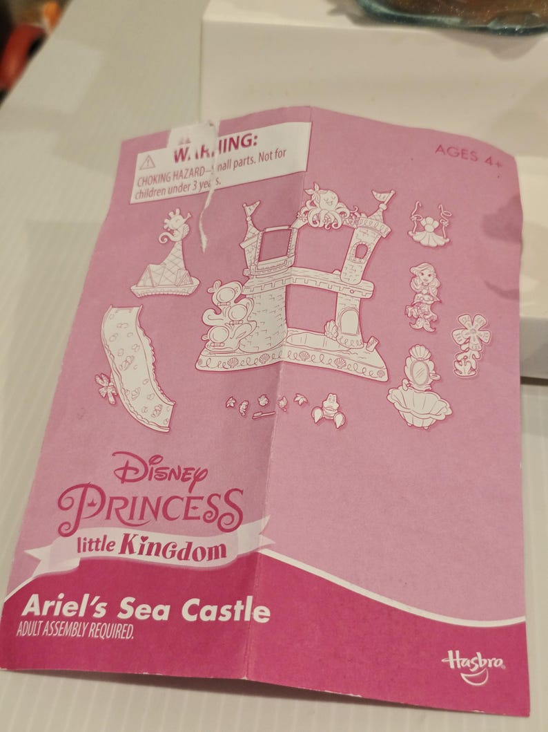 K&ouml;nnte beinhalten: Rosa Anleitung f&uuml;r das Disney Princess Little Kingdom Ariel's Sea Castle Spielset. Die Anleitung zeigt ein Schlossdesign und verschiedene Charakterillustrationen. Der Text enth&auml;lt eine Warnung vor Erstickungsgefahr und das Hasbro-Logo.