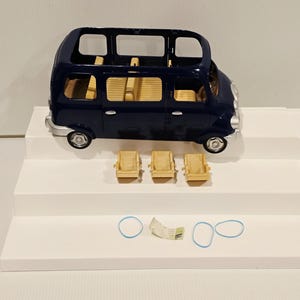 May include: A dark blue toy van with a light tan interior and three matching seats. The van has silver wheels and is sitting on a white surface. Two blue rubber bands and a small paper are also present.