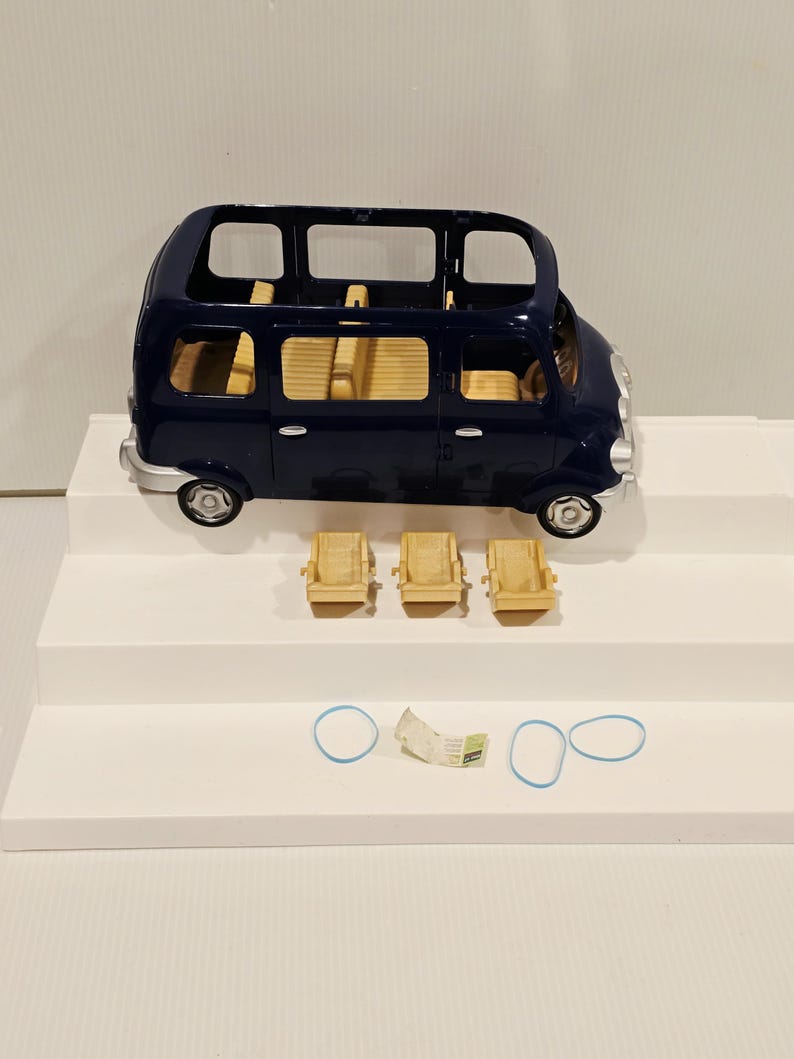 May include: A dark blue toy van with a light tan interior and three matching seats. The van has silver wheels and is sitting on a white surface. Two blue rubber bands and a small paper are also present.