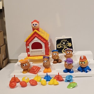 May include: A collection of Mr. Potato Head toys and accessories. The set includes various potato-shaped figures with interchangeable parts, a red and yellow house, and a chalkboard. The accessories include hats, shoes, and other colorful pieces.