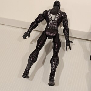May include: Black Spider-Man action figure with a white spider emblem on the back. The figure is in a dynamic pose, showcasing its textured suit and detailed design. The figure is a collectible item.