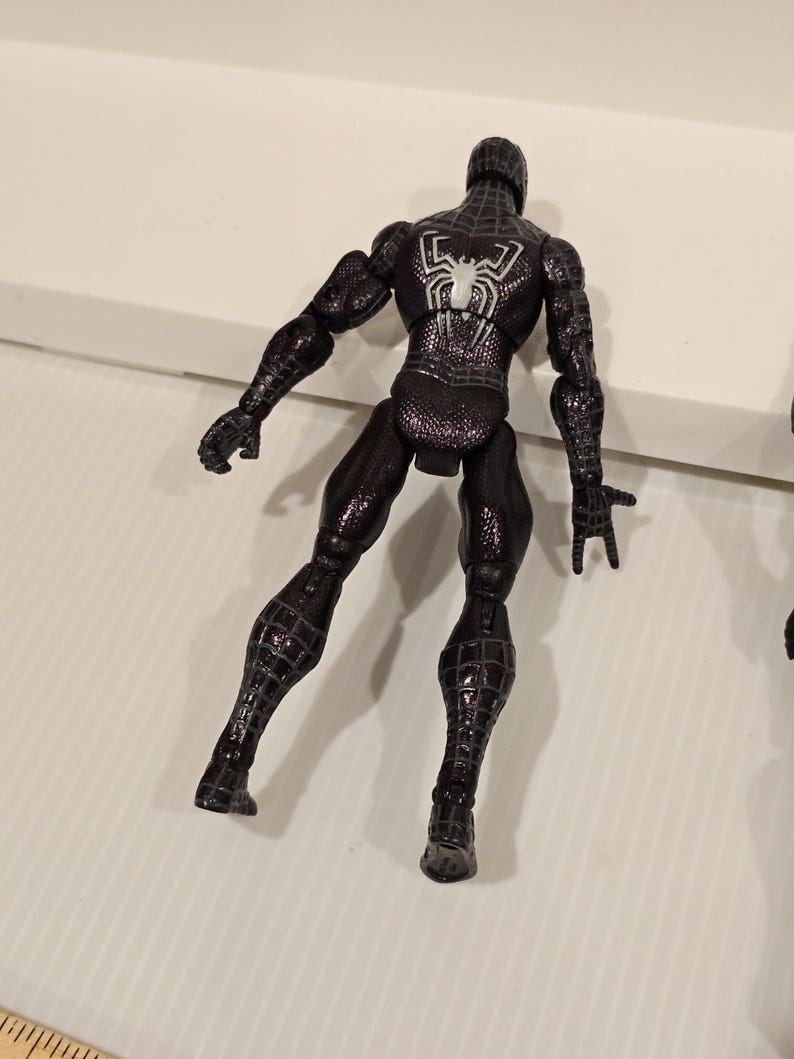 May include: Black Spider-Man action figure with a white spider emblem on the back. The figure is in a dynamic pose, showcasing its textured suit and detailed design. The figure is a collectible item.