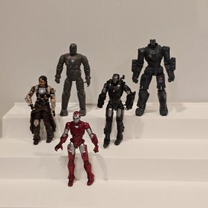 May include: Five action figures of Iron Man characters in various armor designs. The figures are in shades of red, black, and gray. The figures are standing on a white surface.