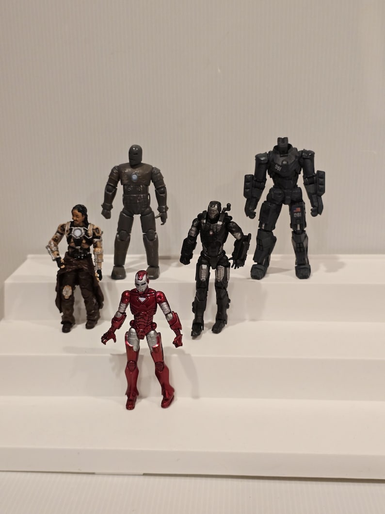 May include: Five action figures of Iron Man characters in various armor designs. The figures are in shades of red, black, and gray. The figures are standing on a white surface.