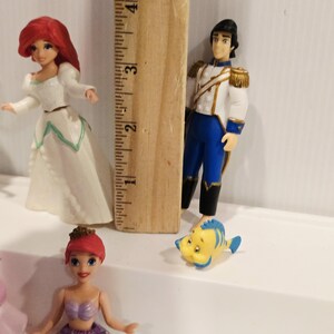 K&ouml;nnte beinhalten: Figuren von Arielle und Prinz Eric, sowie Fabius, aus dem Disney-Film Arielle, die Meerjungfrau. Arielle tr&auml;gt ein wei&szlig;es Kleid und ein lila Oberteil mit gr&uuml;nem Schwanz. Prinz Eric tr&auml;gt eine wei&szlig;e Jacke mit goldenen Akzenten und blaue Hosen.