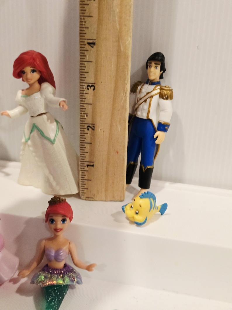K&ouml;nnte beinhalten: Figuren von Arielle und Prinz Eric, sowie Fabius, aus dem Disney-Film Arielle, die Meerjungfrau. Arielle tr&auml;gt ein wei&szlig;es Kleid und ein lila Oberteil mit gr&uuml;nem Schwanz. Prinz Eric tr&auml;gt eine wei&szlig;e Jacke mit goldenen Akzenten und blaue Hosen.