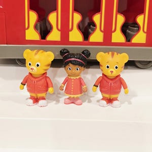 K&ouml;nnte beinhalten: Ein roter Spielzeugzug mit gelben Akzenten und drei Figuren. Zwei Tigerfiguren in roten Jacken und eine M&auml;dchenfigur in einem rosa Kleid stehen vor dem Zug. Die Figuren stehen auf einer wei&szlig;en Oberfl&auml;che.