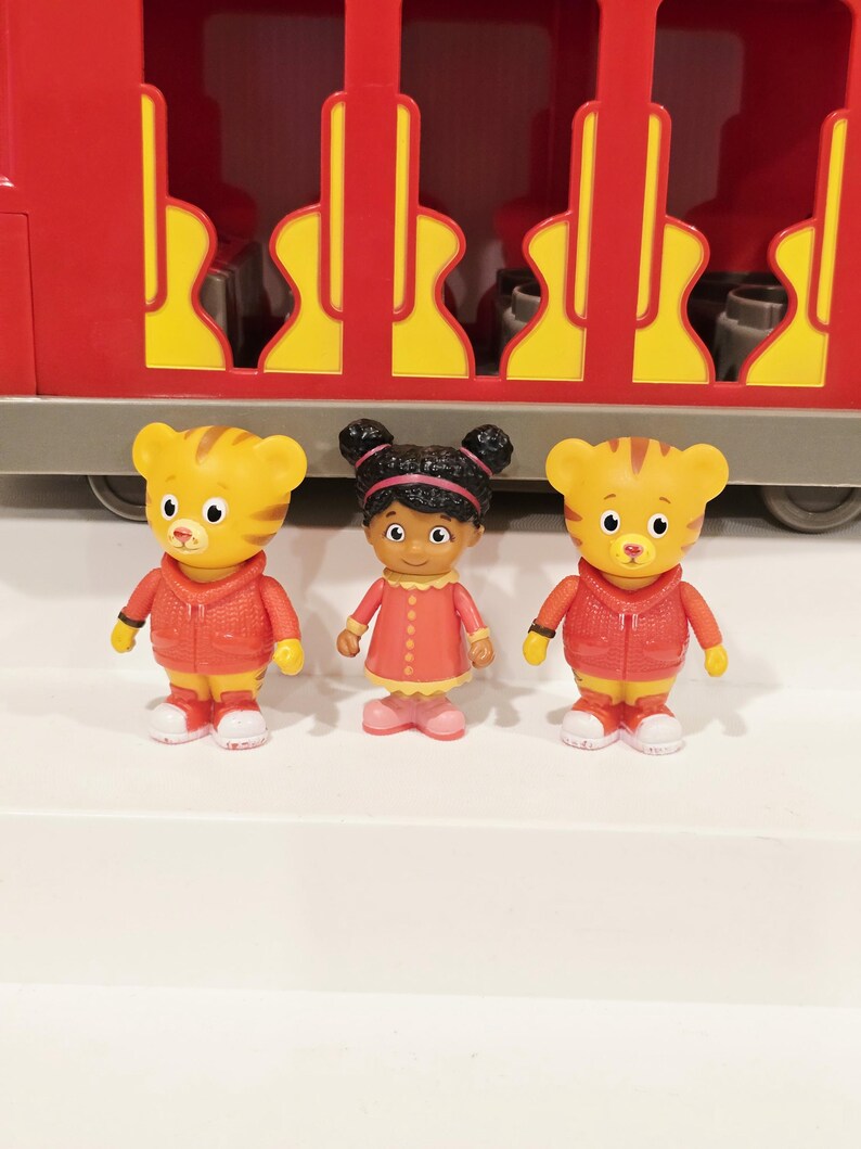 K&ouml;nnte beinhalten: Ein roter Spielzeugzug mit gelben Akzenten und drei Figuren. Zwei Tigerfiguren in roten Jacken und eine M&auml;dchenfigur in einem rosa Kleid stehen vor dem Zug. Die Figuren stehen auf einer wei&szlig;en Oberfl&auml;che.