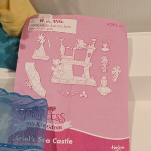 May include: Pink paper with illustrations of a castle, characters, and accessories. The text reads "Ariel's Sea Castle" and "Disney Princess Little Kingdom." The warning label states "Choking hazard - small parts. Not for children under 3 years."