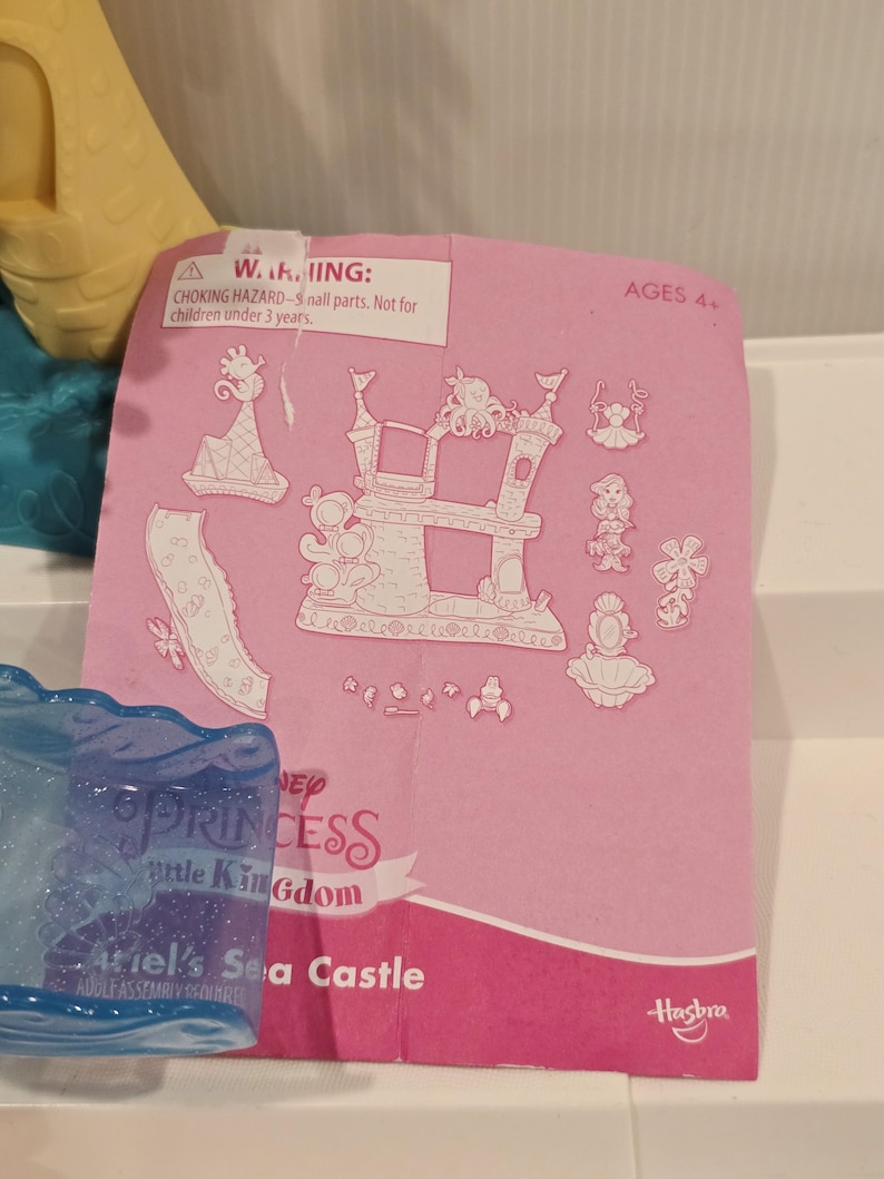 May include: Pink paper with illustrations of a castle, characters, and accessories. The text reads "Ariel's Sea Castle" and "Disney Princess Little Kingdom." The warning label states "Choking hazard - small parts. Not for children under 3 years."