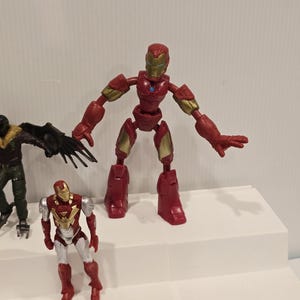 May include: A red and gold Iron Man action figure with articulated joints, standing on a white surface. The figure has a detailed helmet and chest plate. Another Iron Man figure and a Vulture figure are also visible.
