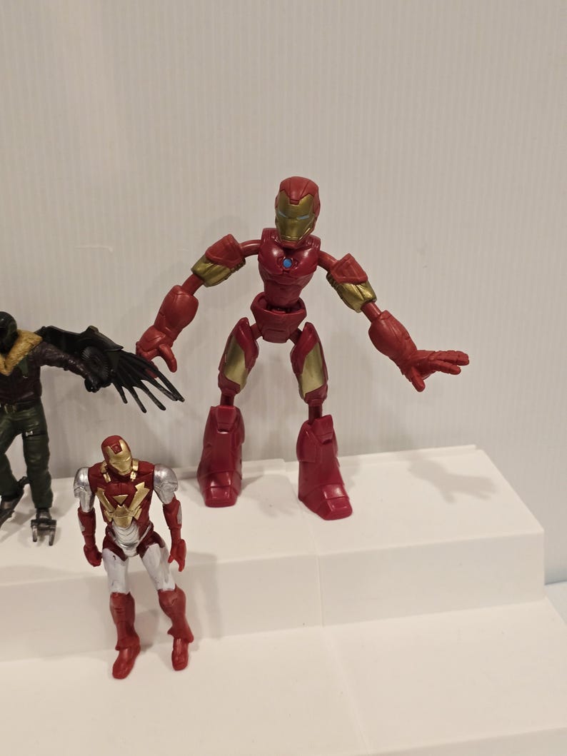 May include: A red and gold Iron Man action figure with articulated joints, standing on a white surface. The figure has a detailed helmet and chest plate. Another Iron Man figure and a Vulture figure are also visible.