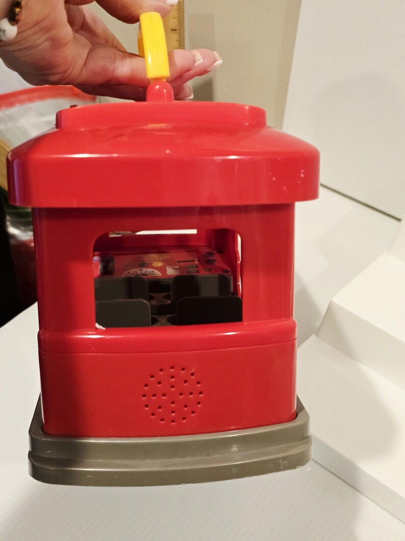 K&ouml;nnte beinhalten: Ein roter Spielzeugbus aus Kunststoff mit einem gelben Griff oben. Der Bus hat einen grauen Sockel und ein Lautsprecher-&auml;hnliches Design an der Seite. Der Bus hat eine rechteckige &Ouml;ffnung in der Mitte mit schwarzen Sitzen.