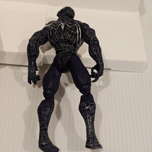 May include: A black Spider-Man action figure, viewed from the back, showcasing intricate web-like detailing. The figure is in a dynamic pose, with articulated joints and a textured suit. The figure's design is a dark color scheme.