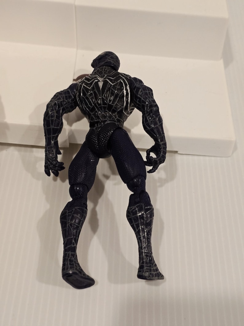 May include: A black Spider-Man action figure, viewed from the back, showcasing intricate web-like detailing. The figure is in a dynamic pose, with articulated joints and a textured suit. The figure's design is a dark color scheme.