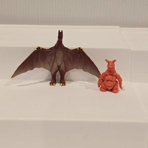 May include: Two toy figures: a brown winged creature with yellow accents and a small orange monster. The winged figure has outstretched wings and stands upright. The orange monster is squatting. Both figures are on a white surface.