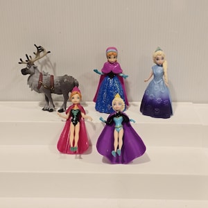 May include: A collection of miniature figurines featuring characters from the animated film "Frozen." The set includes Elsa, Anna, and other characters, with detailed costumes and accessories. The figures are displayed on a white stepped surface.